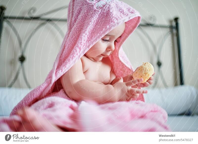 Baby girl in pink towel playing bath sponge while sitting on bed in bedroom color image colour image Spain indoors indoor shot indoor shots interior