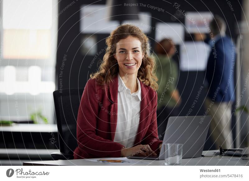 Portrait of smiling businesswoman using laptop at desk in office with colleagues in background Occupation Work job jobs profession professional occupation