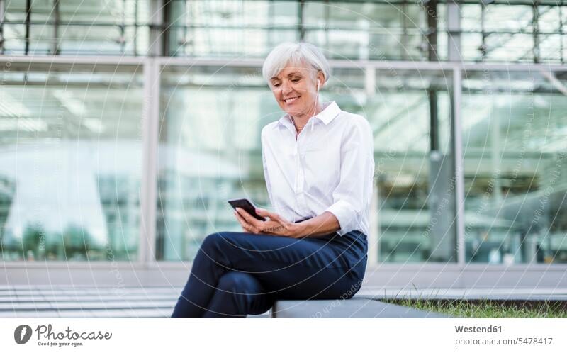 Senior businesswoman sitting outside with smartphone and earbuds businesswomen business woman business women Seated females mobile phone mobiles mobile phones
