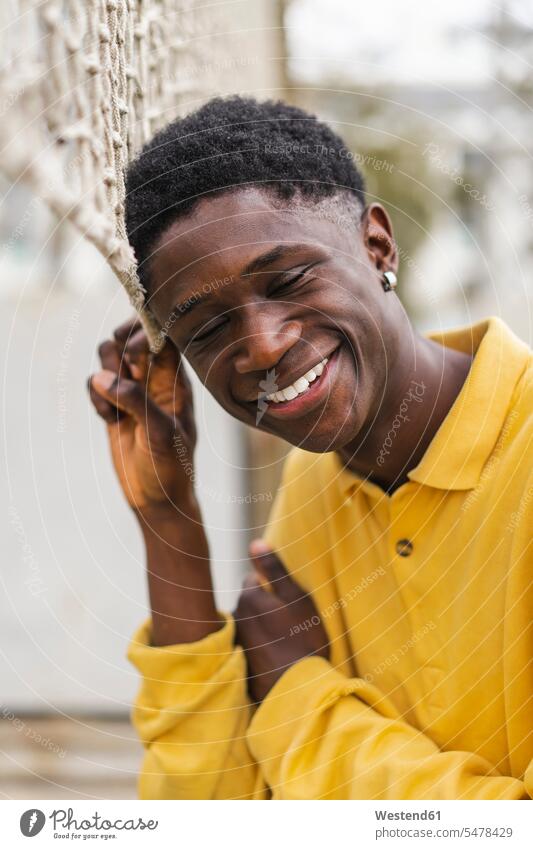 Portrait of a young black man standing by volleyball net, laughing African descent coloured portrait portraits yellow shirt shirts nets young man young men