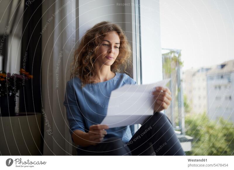 Woman reading paper while sitting on window sill at home color image colour image indoors indoor shot indoor shots interior interior view Interiors day
