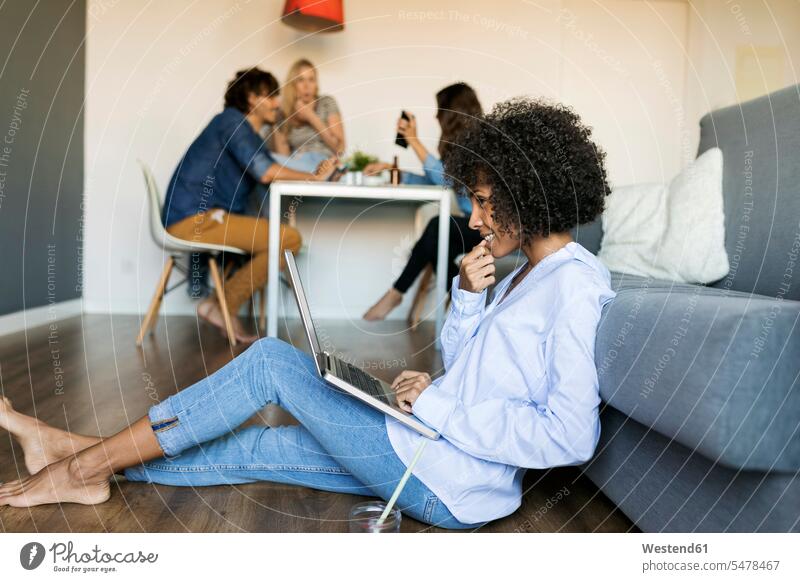 Woman sitting on floor using laptop with friends in background Seated floors woman females women Laptop Computers laptops notebook friendship Adults grown-ups