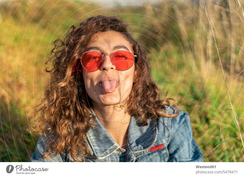 Portrait of young woman with curly brown hair wearing red sunglasses sticking out tongue sun glasses Pair Of Sunglasses females women protruding protrude