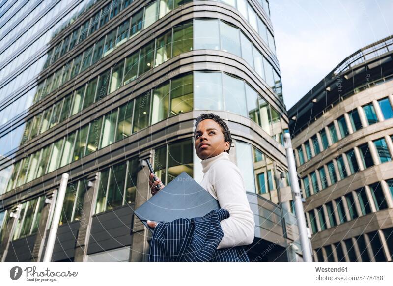 Businesswoman looking away while holding mobile phone and laptop against modern buildings in city color image colour image outdoors location shots outdoor shot