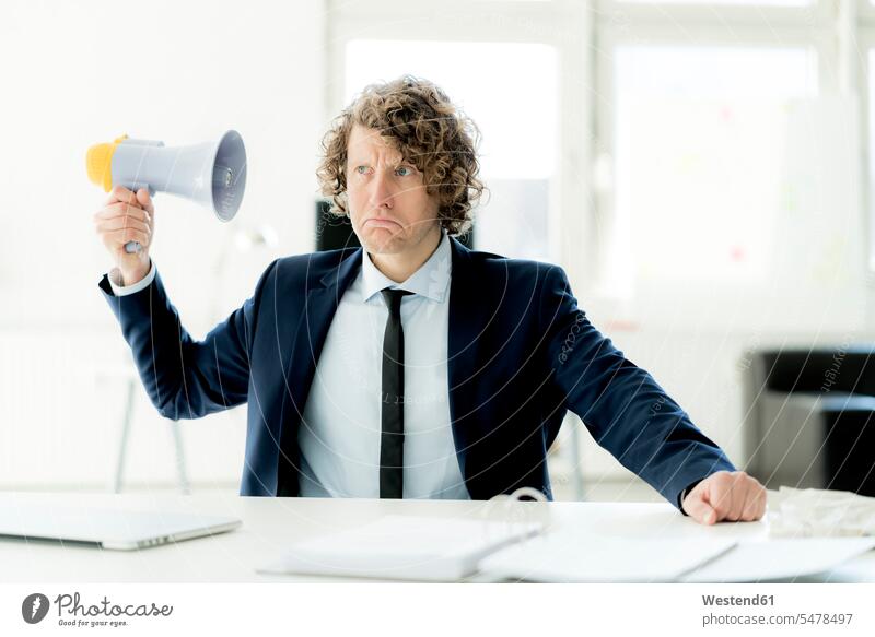 Businessman sitting in office trying out megaphone megaphones bullhorns Discontent discontented unsatisfied dissatisfaction Seated Business man Businessmen