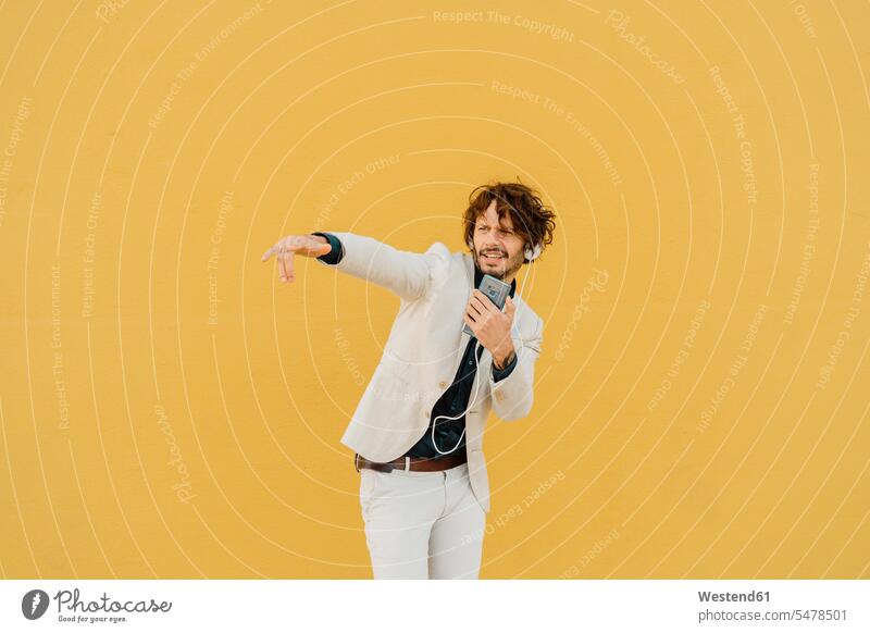 Businessman singing and dancing in front of yellow wall listening music with headphones and smartphone business life business world business person