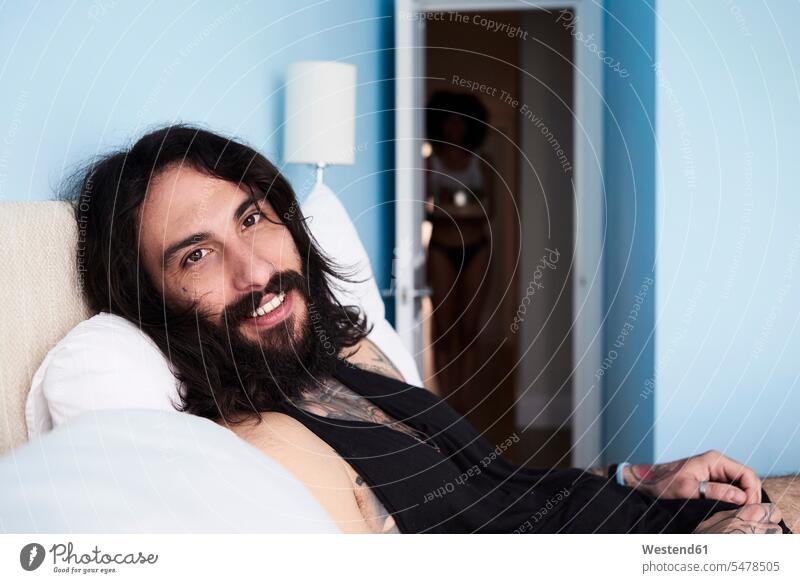 Portrait of smiling tattooed man lying n bed with girlfriend in background portrait portraits beds smile morning in the morning couple twosomes partnership