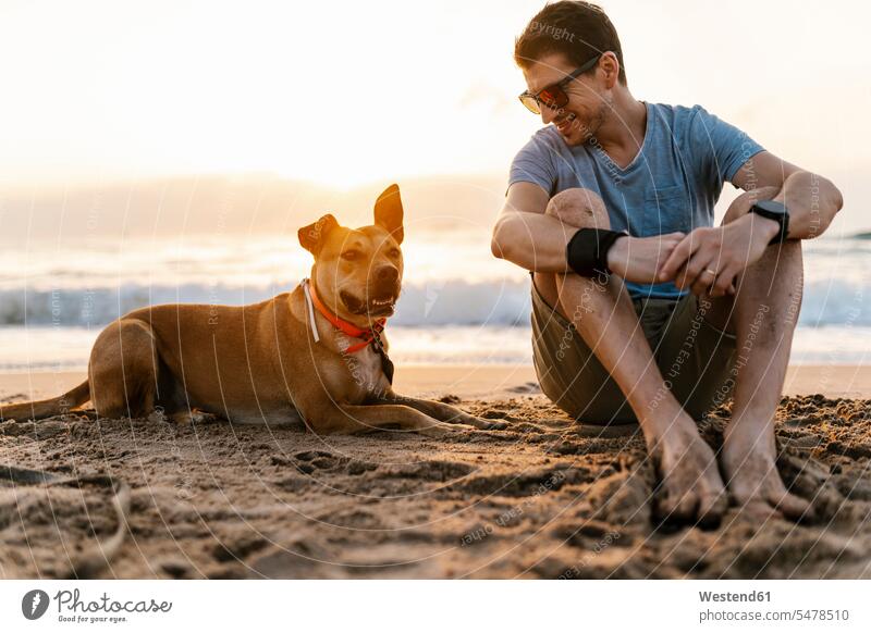 Smiling man looking at his dog while relaxing at beach color image colour image outdoors location shots outdoor shot outdoor shots casual clothing casual wear