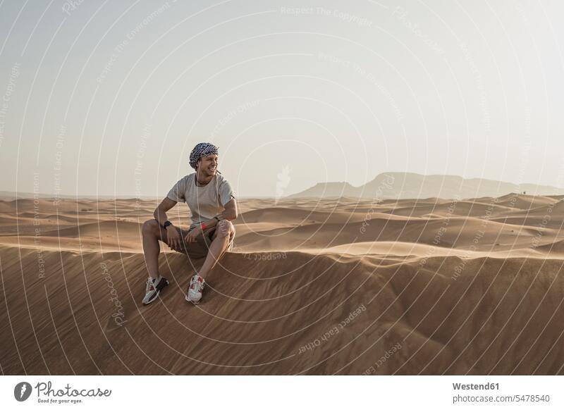 Male tourist looking away while sitting on sand dunes in desert at Dubai, United Arab Emirates color image colour image outdoors location shots outdoor shot