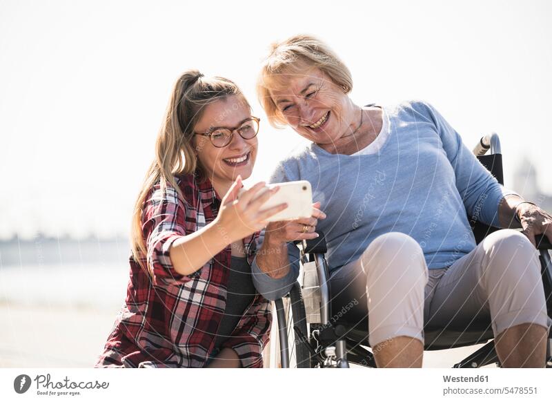 Young woman with her smiling grandmother sitting in wheelchair and taking a selfie generation telecommunication phones telephone telephones cell phone