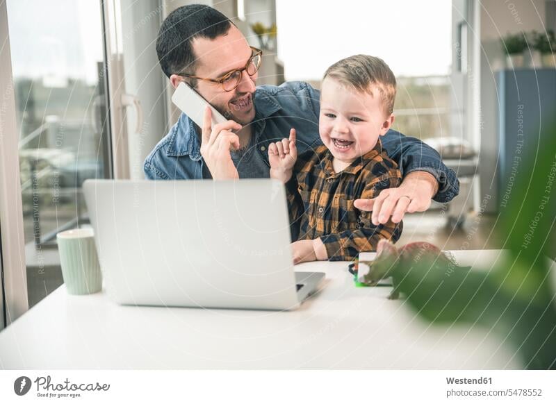 Happy father on cell phone in home office with son sitting on his lap on the phone call telephoning On The Telephone calling working from home home business pa