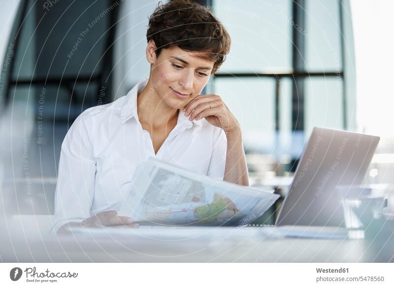 Businesswoman sitting at desk in office with laptop and document desks offices office room office rooms Seated Laptop Computers laptops notebook paper documents
