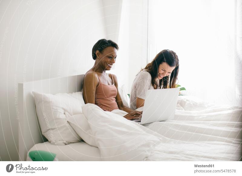 Two happy women lying in bed at home using laptops friends mate female friend human human being human beings humans person persons couples partnership