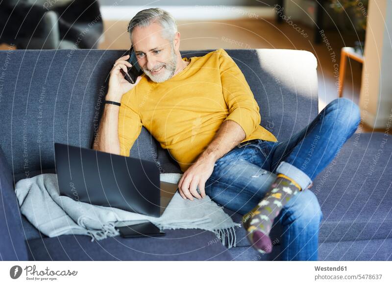 Mature man using cell phone and laptop on couch at home human human being human beings humans person persons celibate celibates singles solitary people