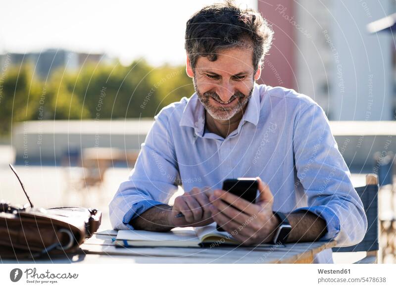 Smiling mature man sitting at outdoor table with cell phone and notebook smiling smile mobile phone mobiles mobile phones Cellphone cell phones men males