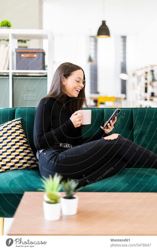 Happy female blogger using phone while sitting with coffee cup on sofa at home color image colour image indoors indoor shot indoor shots interior interior view