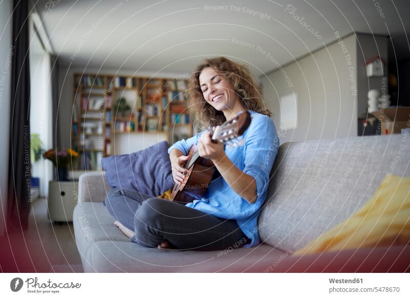 Smiling woman playing guitar while sitting on sofa at home color image colour image indoors indoor shot indoor shots interior interior view Interiors day