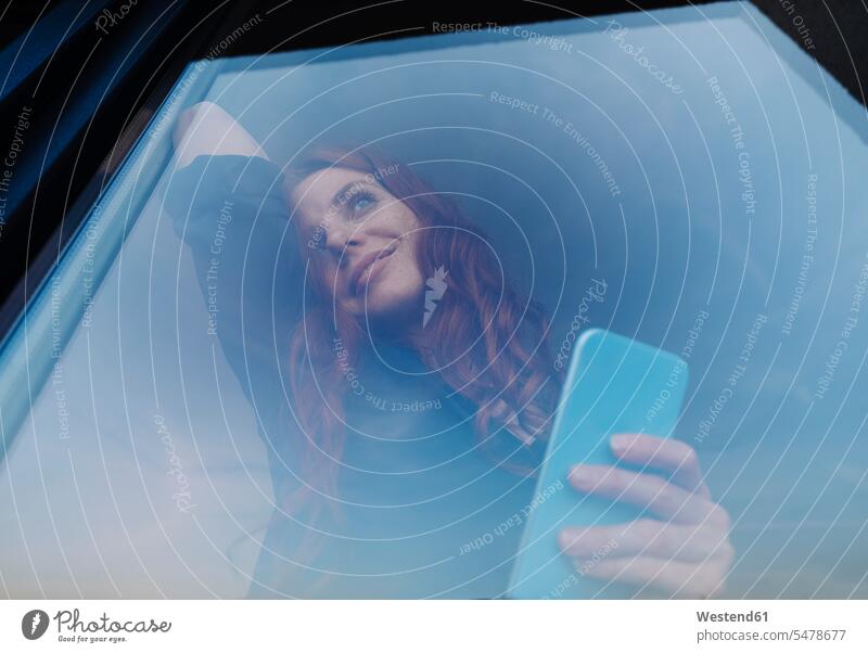 Portrait of smiling redheaded woman holding cell phone behind windowpane business life business world business person businesspeople business woman