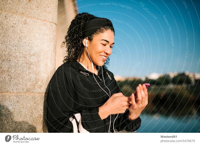 Smiling sportswoman with mobile phone listening music while leaning on column during sunny day color image colour image outdoors location shots outdoor shot