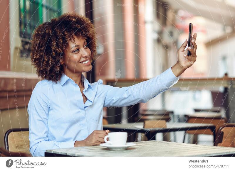 Smiling woman with afro hairstyle sitting in outdoor cafe taking a selfie Selfie Selfies females women Seated smiling smile Afro Afros Adults grown-ups grownups