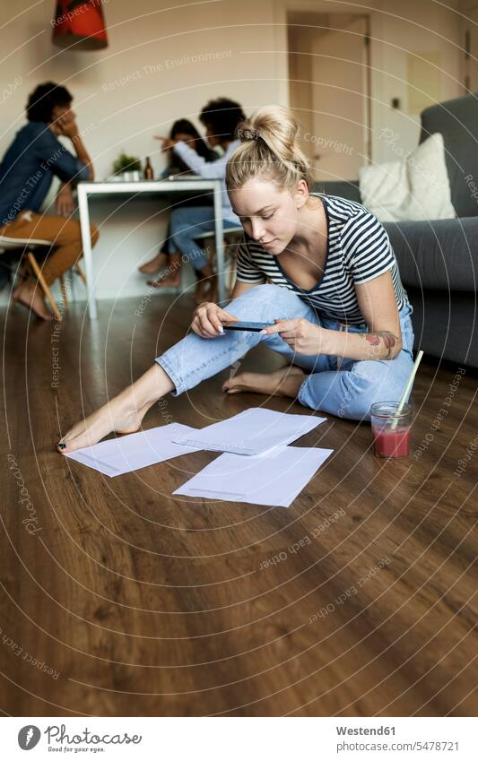 Young woman sitting on floor with cell phone and papers and friends in background floors Seated mobile phone mobiles mobile phones Cellphone cell phones females
