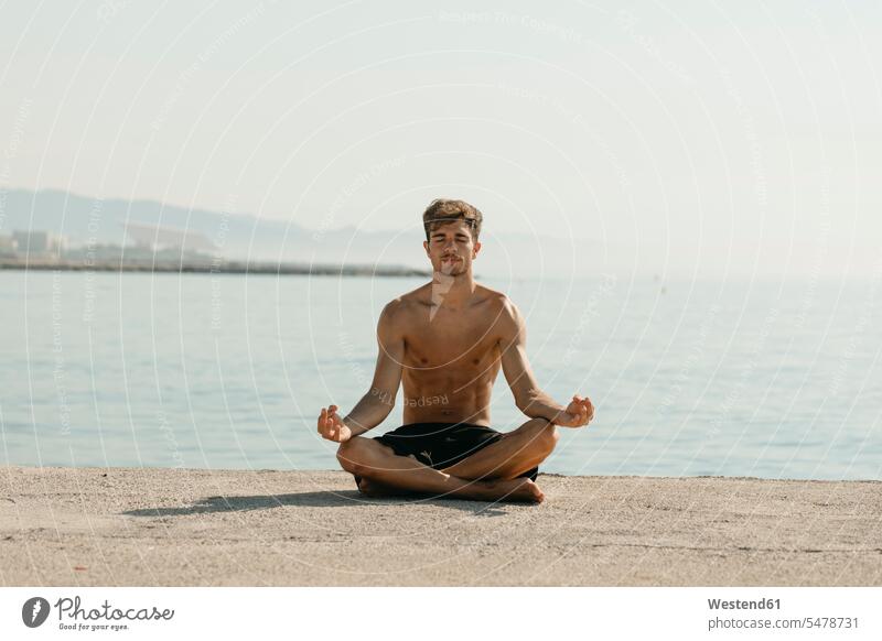 Shirtless young man meditating while sitting against sea at harbor color image colour image outdoors location shots outdoor shot outdoor shots day daylight shot