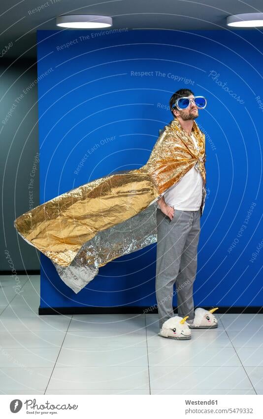 Creative businessman wearing cape made of gold color wrapping paper against blue wall in office color image colour image indoors indoor shot indoor shots