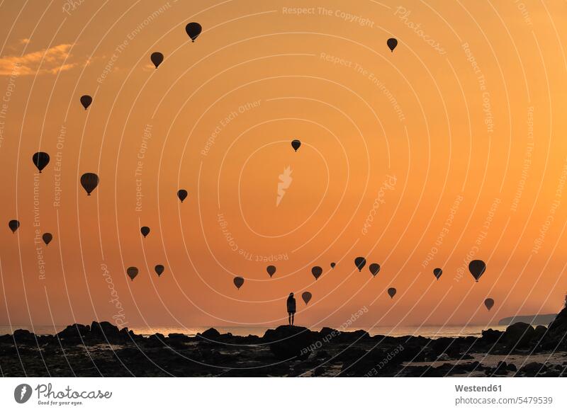 Indonesia, West Nusa Tenggara, Silhouettes of hot air balloons flying over lone woman standing on rocky shore at moody dusk outdoors location shots outdoor shot