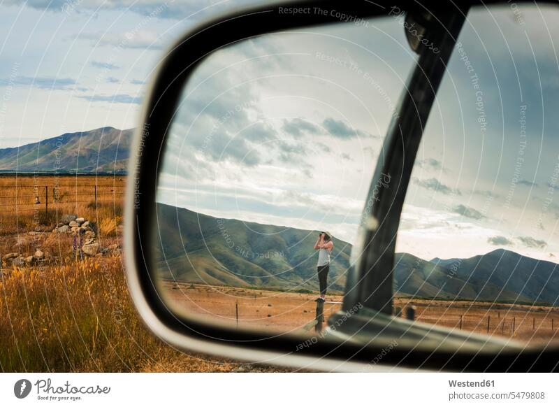 Side-view mirror reflection of young man standing on top of fence looking through binoculars at surrounding mountains outdoors location shots outdoor shot