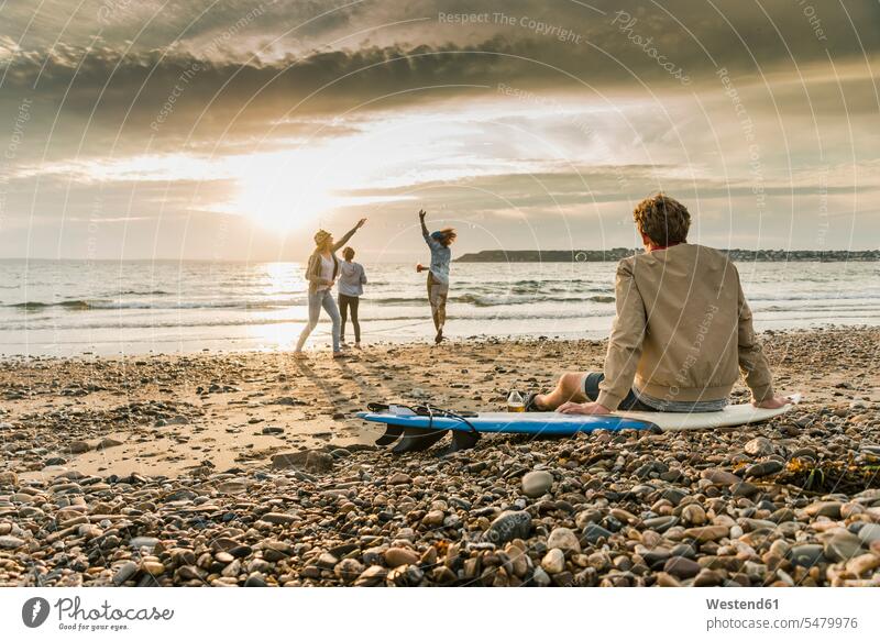 Young man on surfboard watching friends making soap bubbles on the beach at sunset happiness happy relaxed relaxation surfboards beaches relaxing friendship