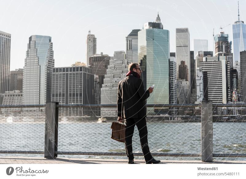 USA, Brooklyn, businessman with briefcase and smartphone standing in front of Manhattan skyline Businessman Business man Businessmen Business men