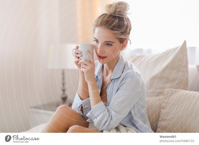 Portrait of daydreaming blond woman sitting on bed with cup of coffee caucasian caucasian ethnicity caucasian appearance european beds indoors indoor shot