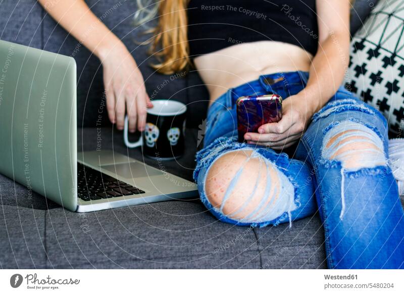 Young woman sitting on couch, using smartphone and laptop cozy sociable comfortable cosy text messaging SMS Text Message reading Seated home at home