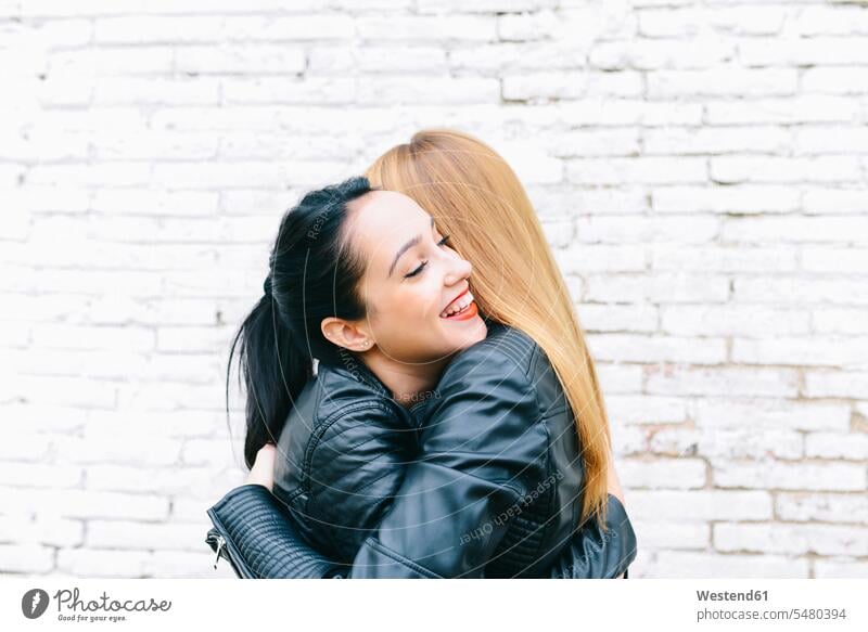 Two young women hugging in front of white brick wall female friends woman females embracing embrace Embracement mate friendship Adults grown-ups grownups adult