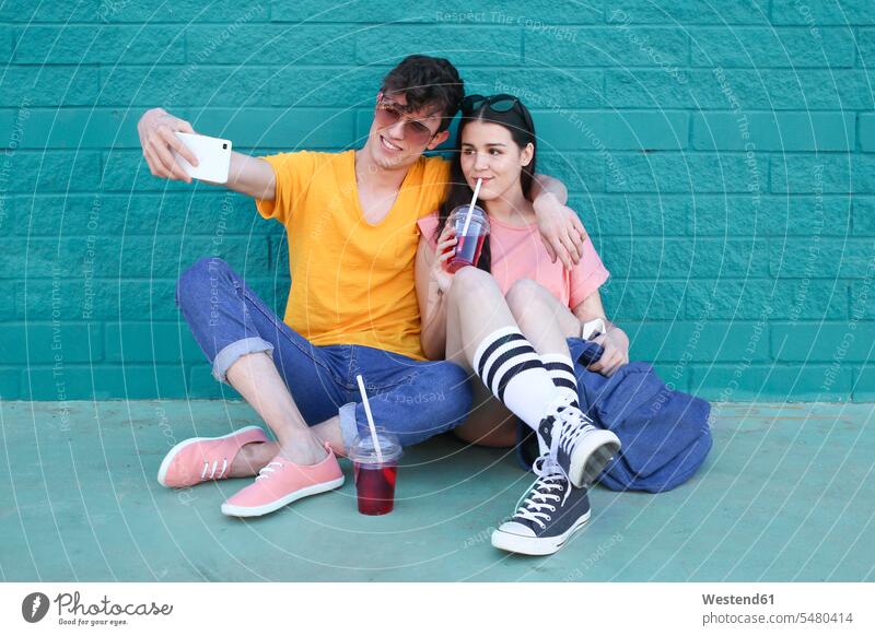 Young couple with soft drinks taking selfie with smartphone in front of blue brick wall Selfie Selfies twosomes partnership couples people persons human being