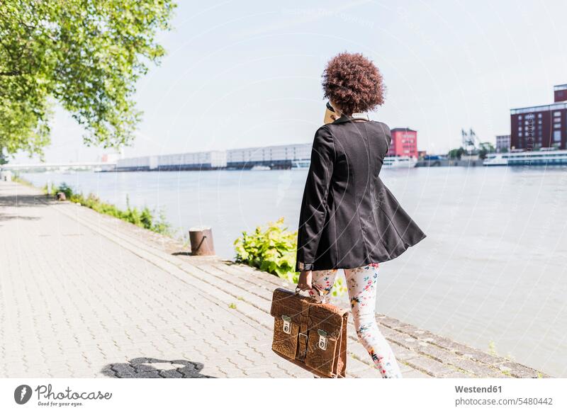 Germany, Mannheim, back view of young businesswoman with briefcase and coffee to go walking at riverside businesswomen business woman business women