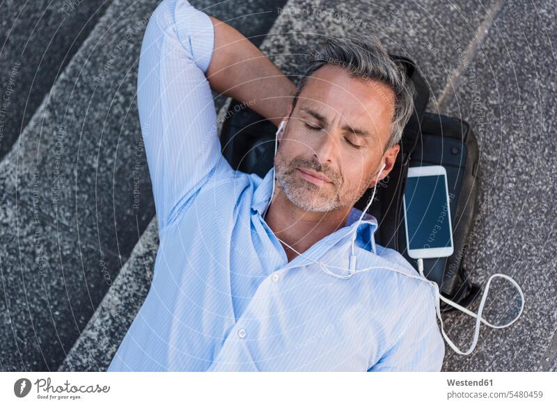 Man with closed eyes lying on stairs with cell phone and earbuds man men males relaxed relaxation laying down lie lying down mobile phone mobiles mobile phones