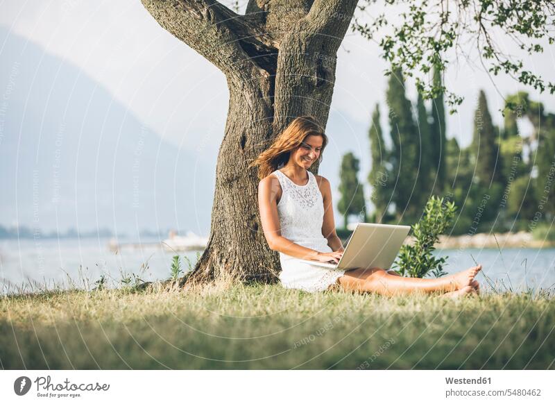 Italy, Lake Garda, young woman leaning against a tree using laptop sitting Seated smiling smile Laptop Computers laptops notebook females women Tree Trees Table