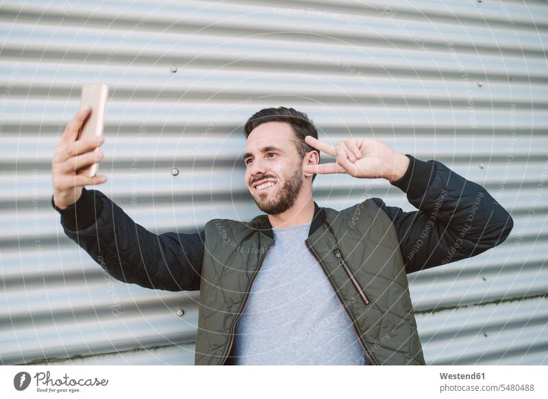 Portrait of smiling man taking selfie with smartphone showing victory sign V sign v-sign men males Selfie Selfies Smartphone iPhone Smartphones smile portrait