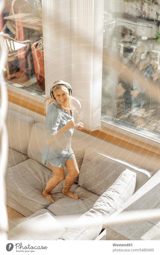Happy woman at home wearing headphones dancing on the couch females women dance settee sofa sofas couches settees headset living room living rooms livingroom