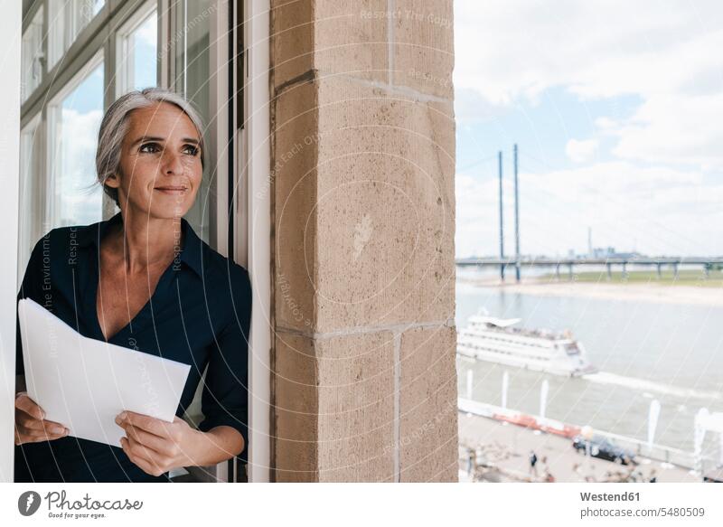Smiling businesswoman looking out of window in waterfront office females women windows smiling smile businesswomen business woman business women Adults