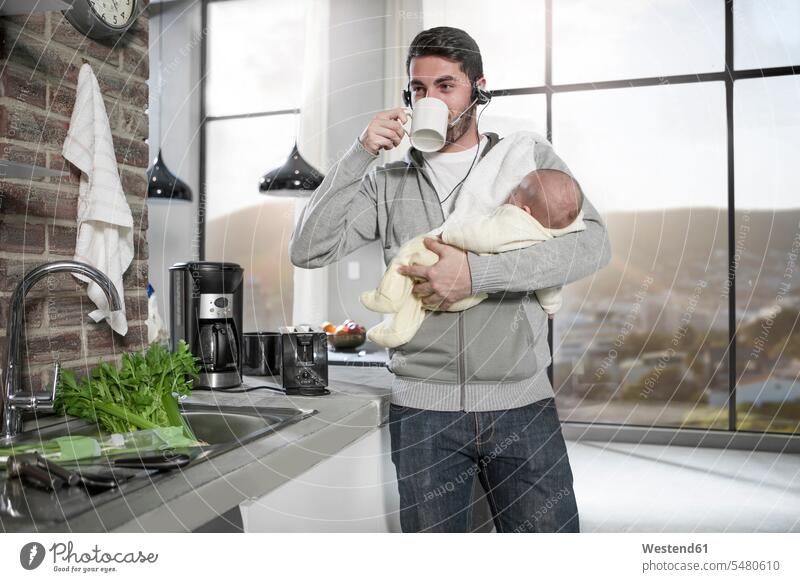 Father with headset drinking coffee in kitchen holding baby on the phone call telephoning On The Telephone calling father pa fathers daddy dads papa