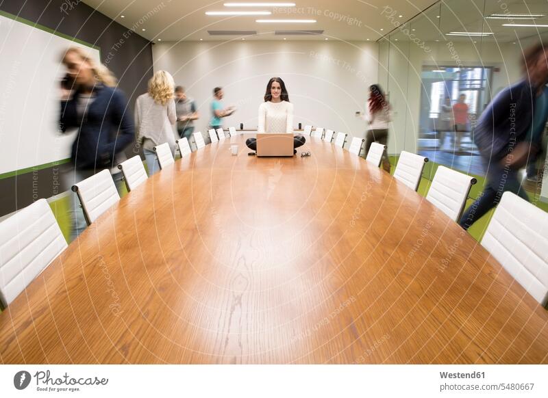 Businesswoman with laptop sitting on conference table with people running around relaxed relaxation Female Colleague Office Offices hustle urgency board room