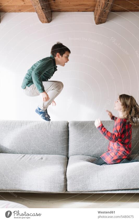 Boy jumping in the air on the couch while his sister watching him settee sofa sofas couches settees sisters Leaping boy boys males jump in the air siblings