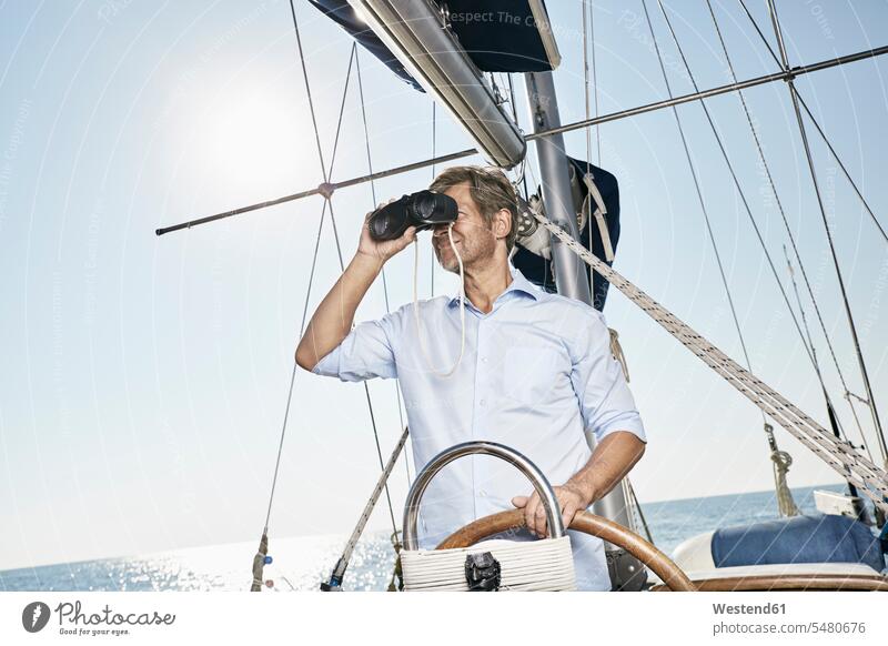 Mature man using binoculars at helm of his sailing boat men males Adults grown-ups grownups adult people persons human being humans human beings boat sports