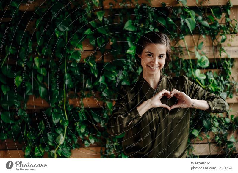 Portait of smiling young woman shaping heart at wall with climbing plants environment ecology environmental females women portrait portraits smile hearts