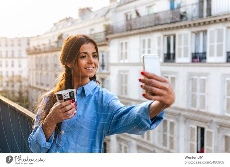 Young woman standing on balcony takinf smart phone selfie females women young using use balconies Coffee Coffee Cup Coffee Cups Selfie Selfies Adults grown-ups