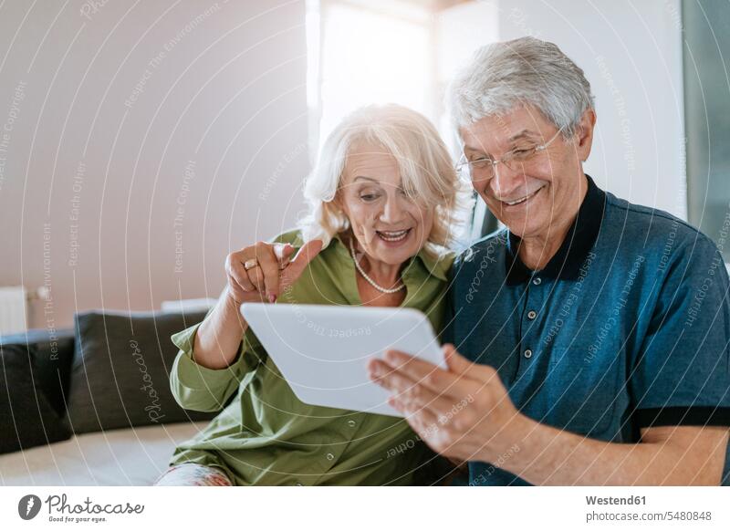 Happy senior couple at home sitting on couch sharing tablet twosomes partnership couples laughing Laughter settee sofa sofas couches settees digitizer