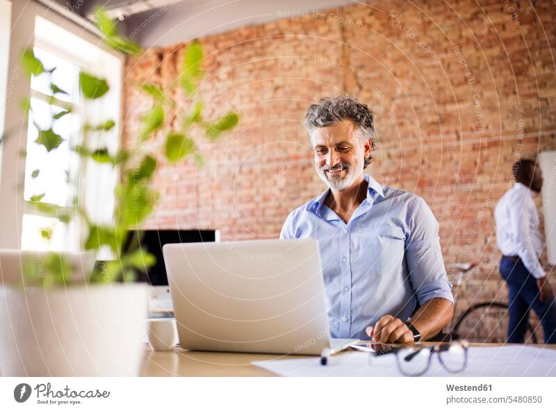 Smiling businessman using laptop in office with colleague in background smiling smile offices office room office rooms Laptop Computers laptops notebook