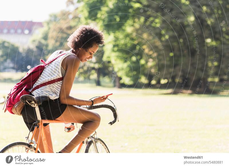 Young woman on bicycle in park checking cell phone bikes bicycles smiling smile parks females women mobile phone mobiles mobile phones Cellphone cell phones